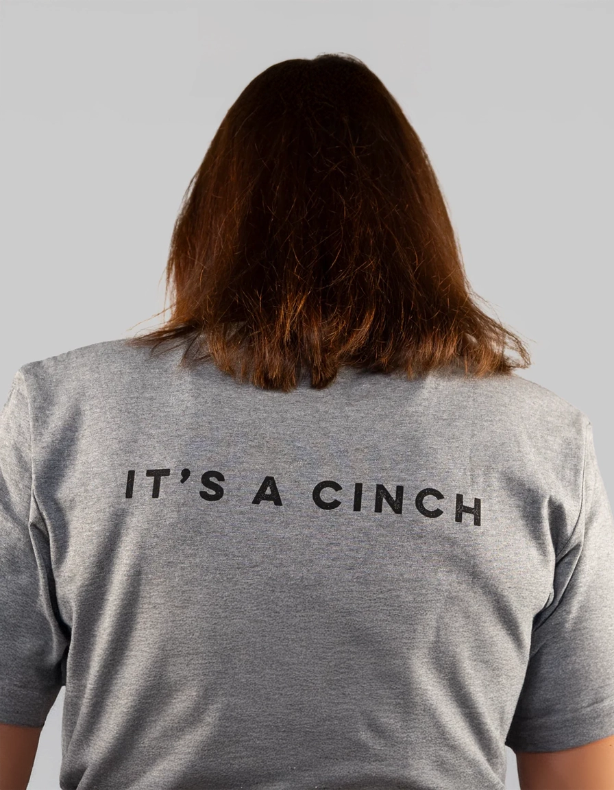 Woman wearing a gray ponlo.com  t-shirt with 'IT'S A CINCH' text on the back against a light gray background