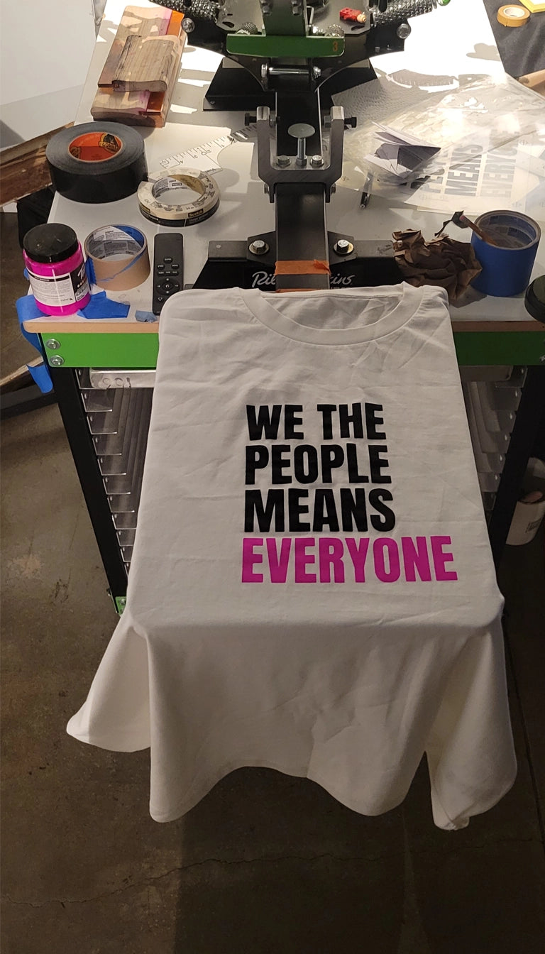 White t-shirt with text 'WE THE PEOPLE MEANS EVERYONE' on a table in a workshop.