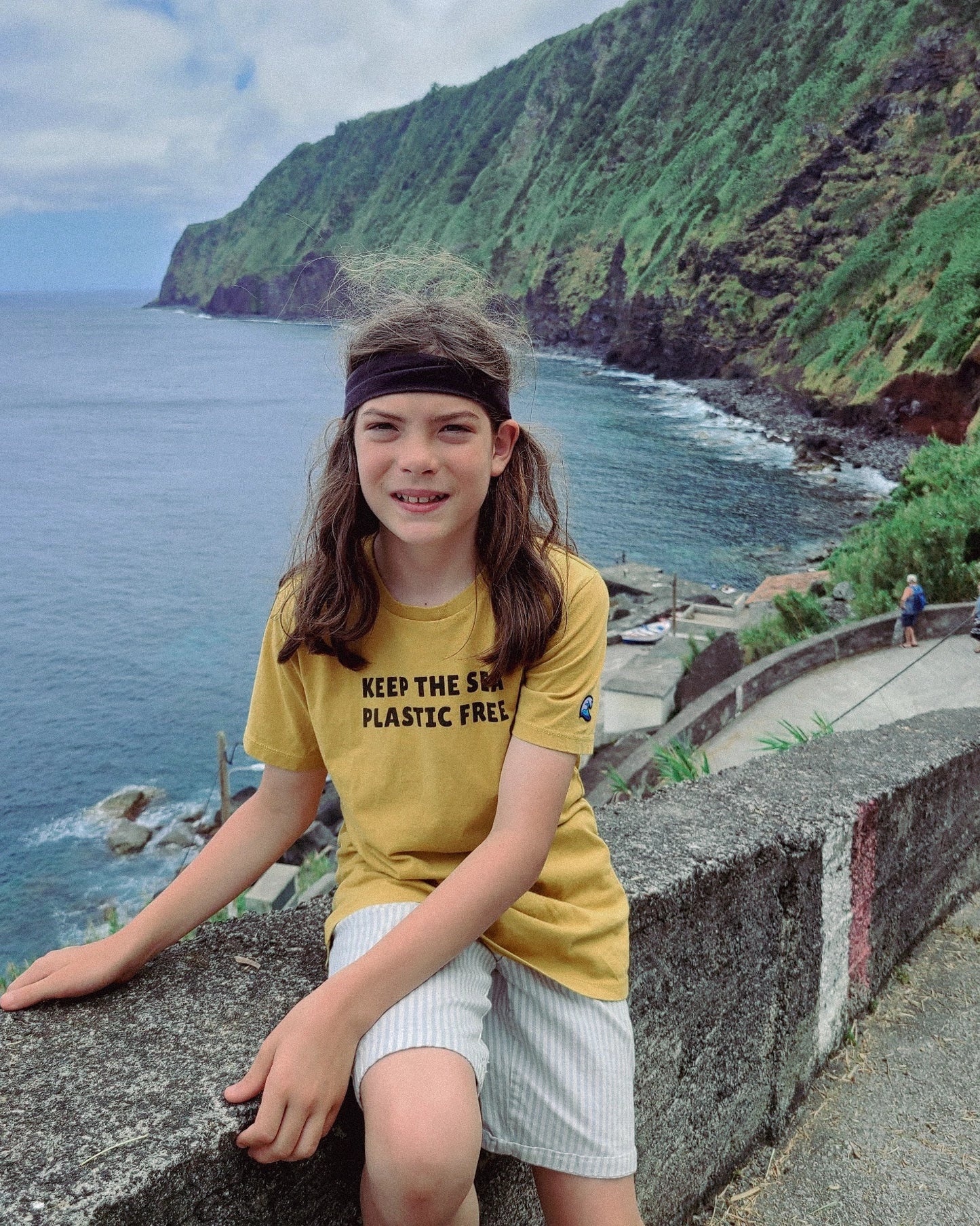 Person wearing a yellow ponlo.com t-shirt with an environmental message, sitting by a coastal landscape.